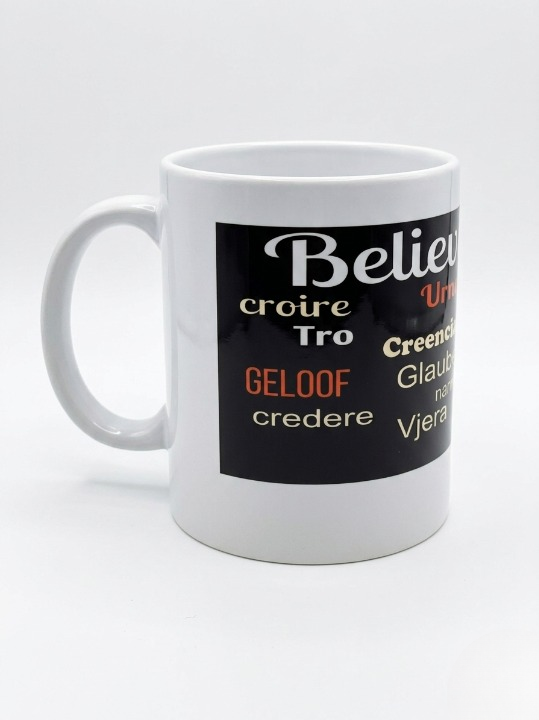 A left side view of handle of white 11-ounce ceramic mug featuring a wide black wrap-around band. The word "Believe!" is prominently displayed at the top in large, white cursive script. Below it, the word is translated into various languages. The design is clean and inspirational, set against a bright, minimalist white background.   Shop www.ANDREAANDME.com
