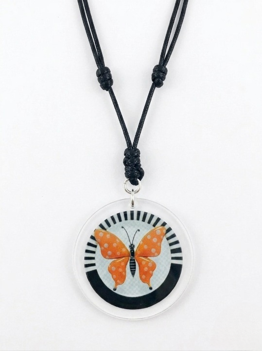 Front view of an orange and white polka dots butterfly acrylic and mixed media pendant necklace. The graphic features a close-up of the circular 2 1/2 inch pendant containing an orange butterfly with white polka dots against a black and white striped background.  Shop at www.andreaandme.com